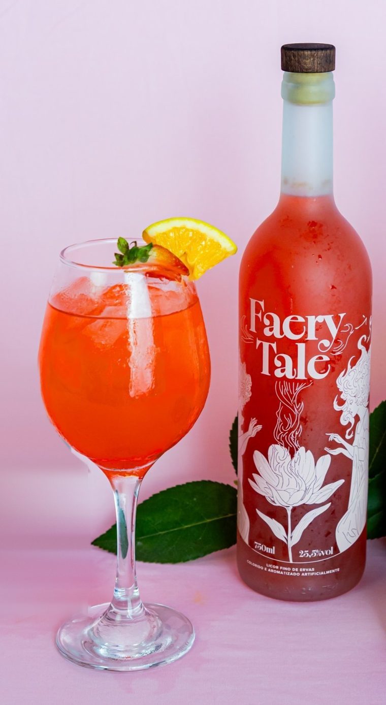 Taça de Faery Spritz com gelo, morango e rodela de laranja ao lado da garrafa de Faery Tale, sobre fundo rosa